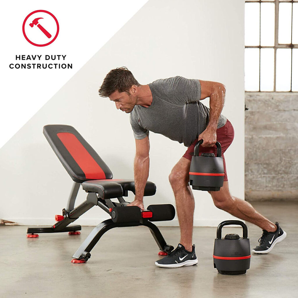 A man lifting a heavy adjustable kettlebell in a home gym, next to an exercise bench labeled "Heavy Duty Construction".