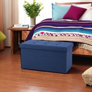 A rectangular navy blue fabric storage ottoman with a button-tufted lid, placed at the foot of a bed on a wooden floor in a modern bedroom
