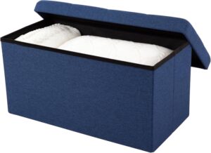 An open navy blue linen-textured storage ottoman showing white folded blankets stored inside. The tufted lid is partially open against a white background.