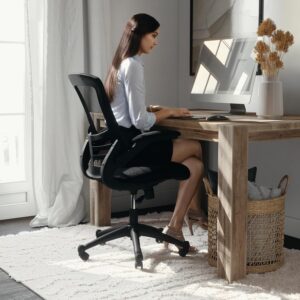 A lifestyle image showing a woman with long hair sitting on a black ergonomic mesh office chair, typing at a computer on a rustic wooden desk. The workspace is styled with a textured white area rug, a woven storage basket, and natural light streaming from a nearby window.